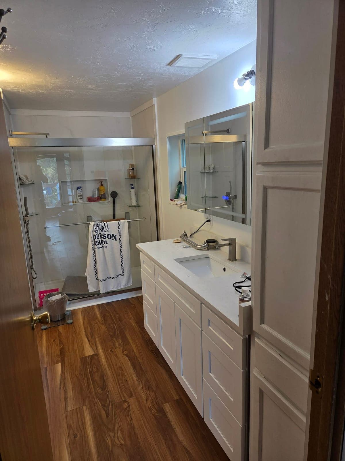 Remodeled bathroom with a white shaker vanity, quartz countertop, wide mirror, sliding glass shower door, and wood-look plank flooring